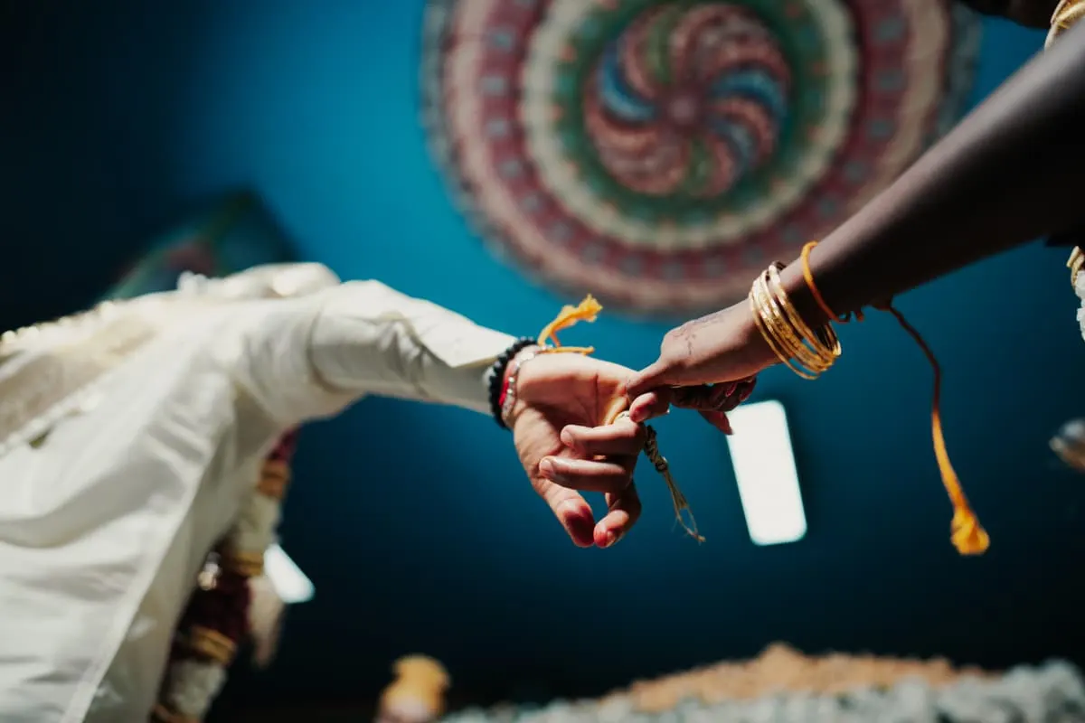 Bride and groom's hands meeting under a painted mandap ceiling, her wrist stacked with gold bangles.