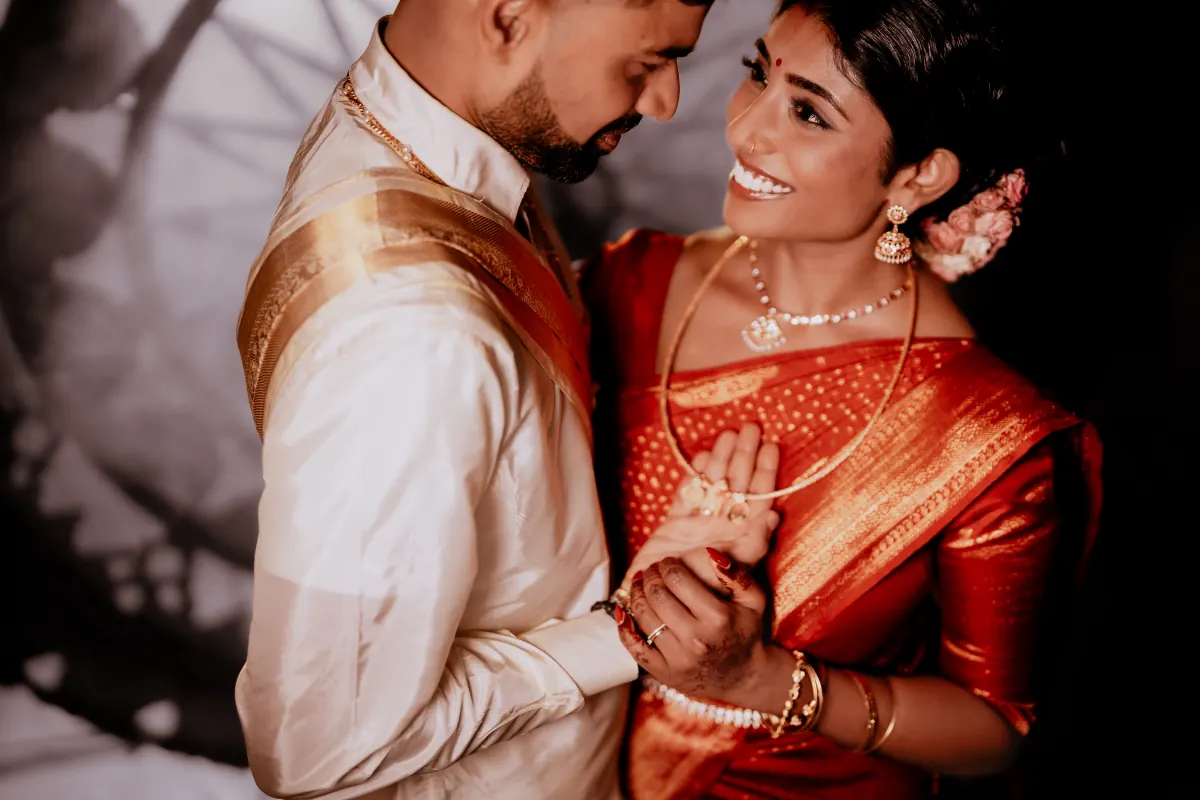 A bride in a red silk sari laughing up at her groom, his gold-bordered veshti over one shoulder.