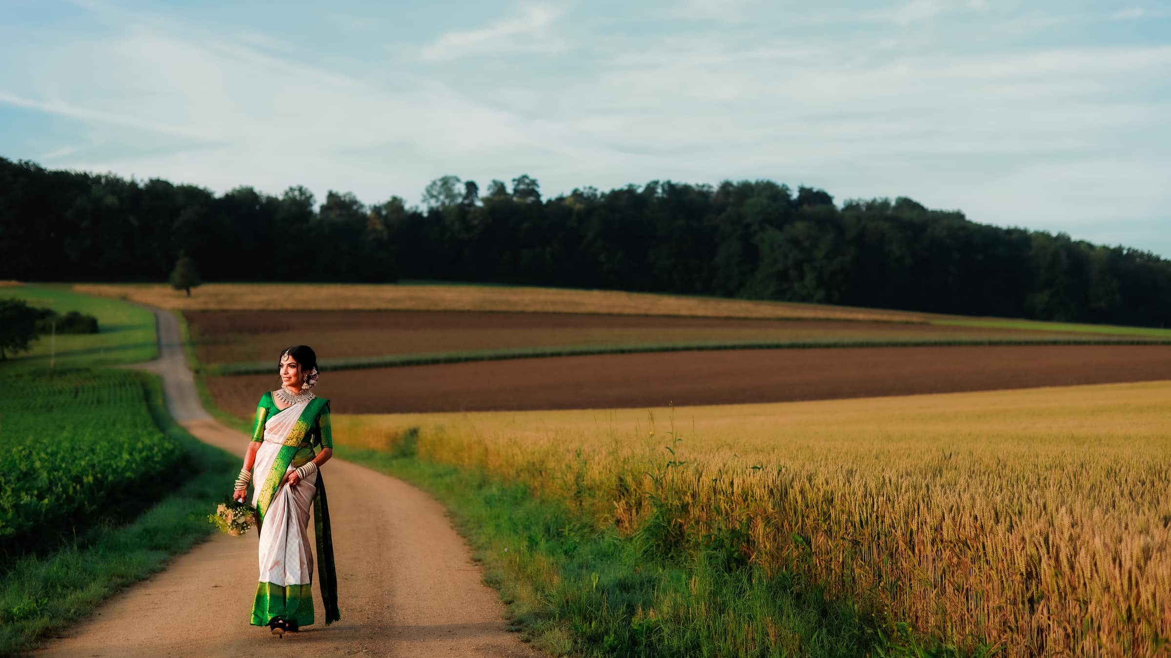 A bride in a cream and green silk sari walking a country road between wheat fields in evening light.